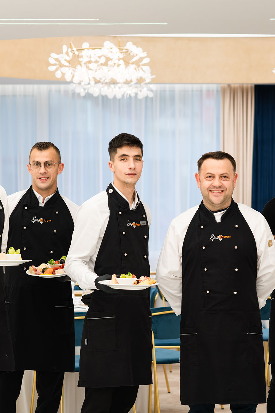 A group of men in black aprons holding plates of food in a hotel kitchen.